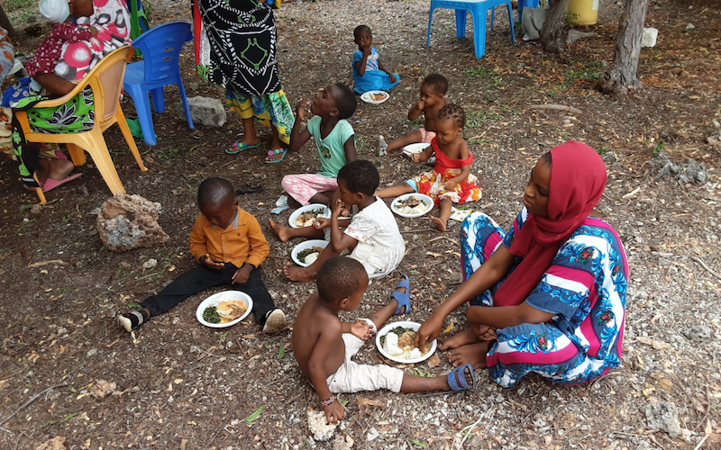 Children and a woman sit on the ground eating from plates